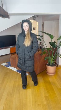 Casaco acolchoado preto com capuz, com bolsos frontais e um design matelassê, usado sobre calças escuras. Chão de madeira e uma planta ao fundo.