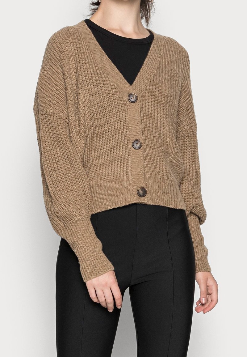 Cardigan marrón de punto con escote en V, que presenta tres botones frontales y hombros caídos, combinado con pantalones ajustados negros.