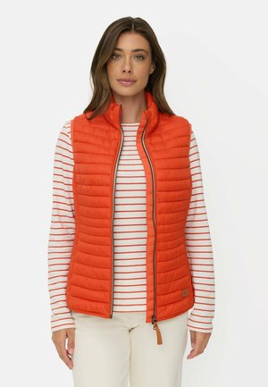 Femme portant un gilet matelassé orange par-dessus une chemise blanche à manches longues avec des rayures orange et un pantalon de couleur claire, debout devant un fond uni.
