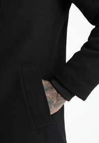Schwarzer Wollmantel mit schlichtem Design, seitlicher Tasche, gerippten Bündchen und weicher Textur. Der Ärmel zeigt aufwendige Tattoos.