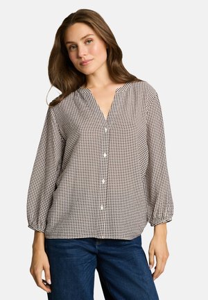 Femme aux longs cheveux bruns portant un chemisier noir à pois blancs avec des boutons et un jean bleu, debout devant un fond uni.