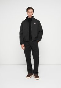 Sort bomberjakke med lynlås foran, ribbede mansjetter og et lille logo. Sammen med mørke bukser og brune sneakers med hvide såler.