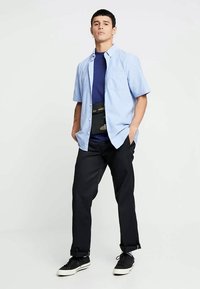 Short-sleeve light blue button-up shirt over a navy t-shirt, paired with black pants and black sneakers, featuring a camo-print waist bag.