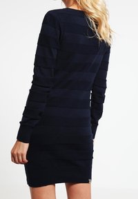 Marinrandig stickad klänning, långa ärmar, figurnära design, ribbad textur, har en logotypdetalj nära fållen och rund halsringning.