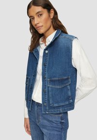 Donkerblauwe denimvest met een kraag, knoopsluiting aan de voorkant en twee voorzakken. Draagt over een wit shirt. Glad textuur.