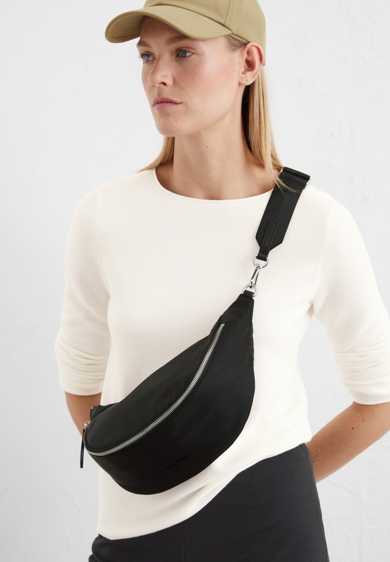 Czarna nylonowa torba na ramię o zakrzywionym kształcie, z ciekawym suwakiem w kolorze srebrnym i regulowanym paskiem, noszona na białej bluzce. Minimalistyczny design z czystymi liniami.