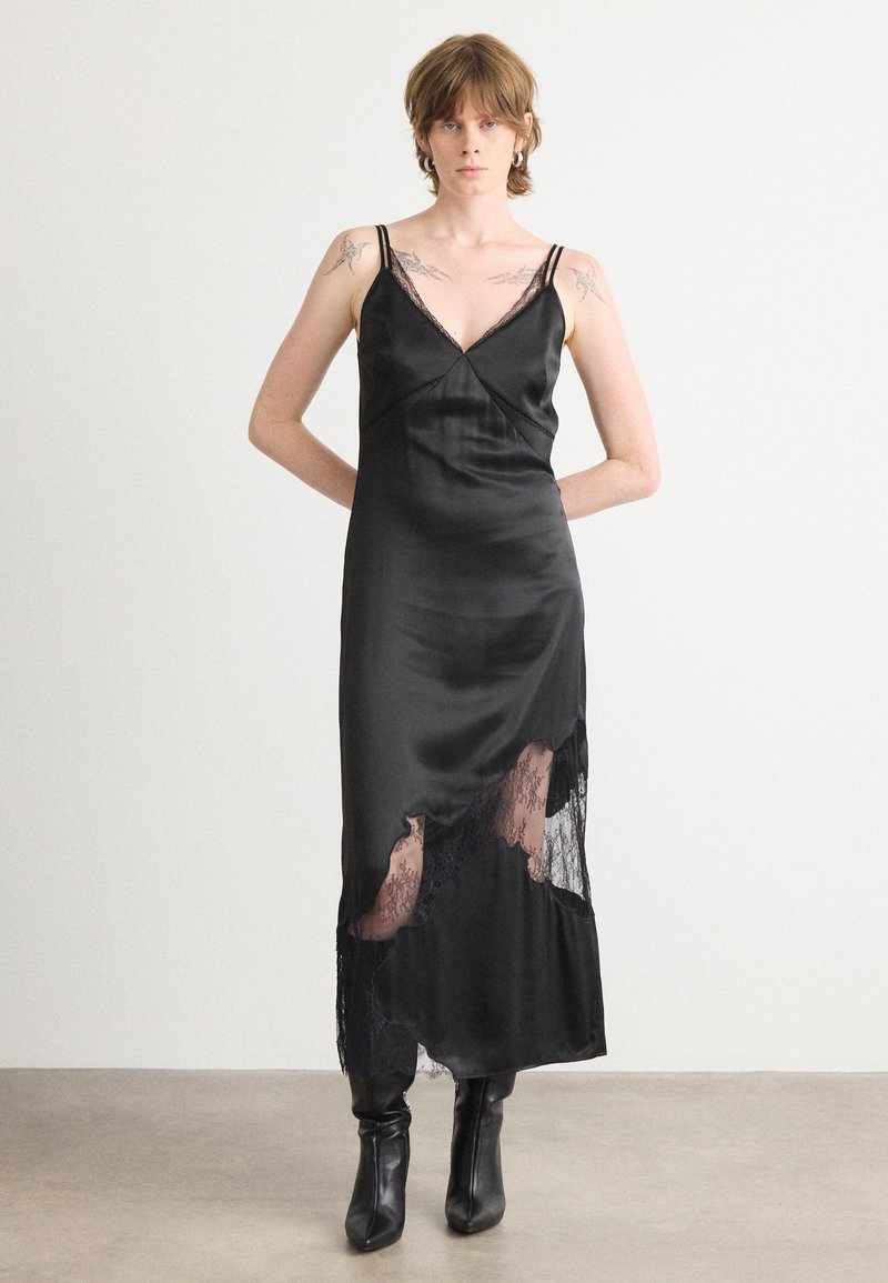 Schwarzes Satin-R slipskleid mit Spitzenakzenten, V-Ausschnitt, dünnen Trägern und asymmetrischem Saum, der ein Spitzenpanel enthüllt. Stiefel runden den Look ab.