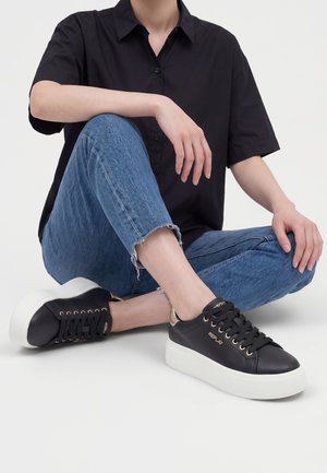 Osoba ubrana w czarną koszulkę z krótkim rękawem, niebieskie dżinsy z postrzępionym dołem oraz czarne sneakersy na platformie z białą podeszwą, siedząca ze skrzyżowanymi nogami na białej podłodze.