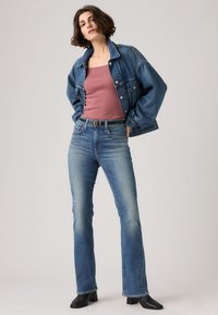 Γυναίκα που φοράει τζιν μπουφάν, ροζ αμάνικη μπλούζα σε χρώμα σκόνης τριαντάφυλλου, φθαρμένο τζιν με σκέλη bootcut, μαύρες μποτάκια αστραγάλου, στέκεται με τα χέρια στις τσέπες μπροστά σε απλό φόντο.