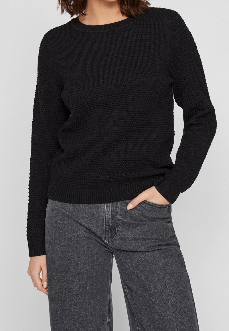 Femme portant un pull en tricot texturé noir et un jean noir délavé, avec une main dans la poche, sur un fond uni.