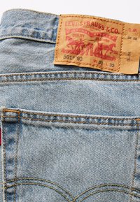 Ljusblå denimjeans med en läderetikett på midjebandet, med tryckt röd text och gul sömnad. Modell 501, storlek W32 L32.