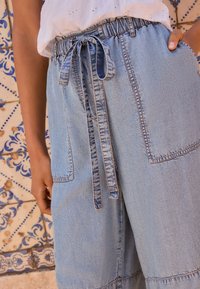 Pantaloni di denim azzurro chiaro con vita elasticizzata e coulisse annodata, dotati di due grandi tasche e dettagli di cuciture visibili.