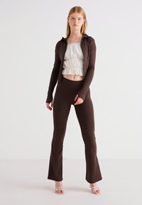 Smešano smeđi zip-up jakna, bijeli naborani crop top i smeđi zvonasti tajice. Model stoji u sandalama s remenčićima na jednostavnoj pozadini.