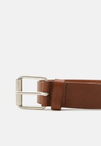 Ceinture en cuir marron avec une boucle en métal argenté et un logo embossé sur la sangle. Texture lisse, forme rectangulaire et design simple.