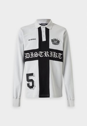 Jeune homme portant un maillot de rugby blanc avec une grande croix noire et un pantalon cargo noir, debout devant un fond blanc uni.