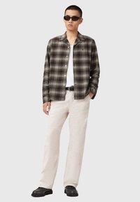 Plaid button-up shirt in dark green and brown, paired with cream-colored corduroy pants and black shoes. Sunglasses worn, casual look.