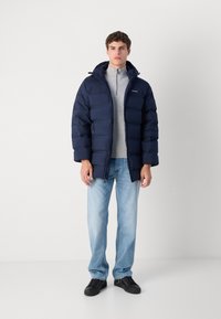 Námornícky puffový kabát, prešívaný dizajn, predný zips, nastaviteľná kapucňa, nosený cez šedý polovičný zips sveter, spárovaný so svetlými džínsami a čiernymi topánkami.