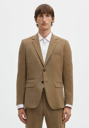 Hombre con cabello castaño que lleva una chaqueta de traje color beige y pantalones a juego sobre una camisa blanca abotonada, de pie frente a un fondo blanco liso.