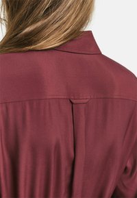 Bordeauxfarbige Bluse mit glatter Textur, ausgestattet mit einem Kragen und einer Faltendetail an der Rückseite, entworfen in einer lässigen Passform.