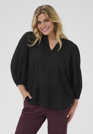 Schwarze Bluse mit V-Ausschnitt, strukturiertem Stoff und Puffärmeln. Kombiniert mit kastanienbraunen Hosen, die eine lockere Passform und subtilen Mustern zeigen.