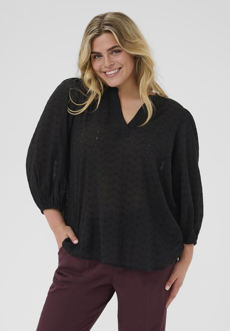 Blouse noire avec un col en V, tissu texturé et manches bouffantes. Associée à un pantalon bordeaux, mettant en valeur une coupe décontractée et des motifs subtils.