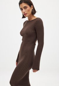 Robe en tricot côtelé marron avec des manches longues, présentant des découpes sur les côtés et une silhouette ajustée. Le tissu texturé crée un motif de rayures verticales.