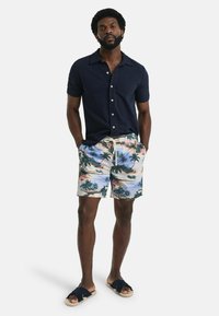 Marineblauwe shirt met korte mouwen, geblokte strandshorts met palmbladeren en zwarte sandalen met zachte zolen. Informele zomerkleding tentoongesteld.