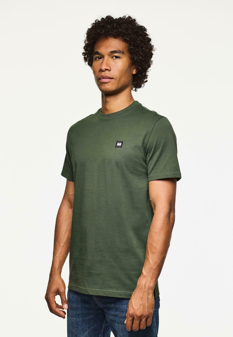 Hombre con cabello rizado, vistiendo una camiseta de manga corta verde lisa y jeans azules, de pie contra un fondo blanco.