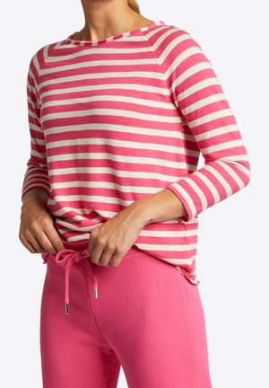 Femme ajustant un pantalon à cordon rose, portant une chemise à manches longues rayée rose et blanche sur un fond uni.