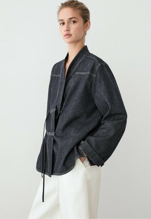 Ung kvinde iført oversized mørk denimjakke med bindelukninger og hvide bukser, stående med den ene hånd i lommen, neutral baggrund.