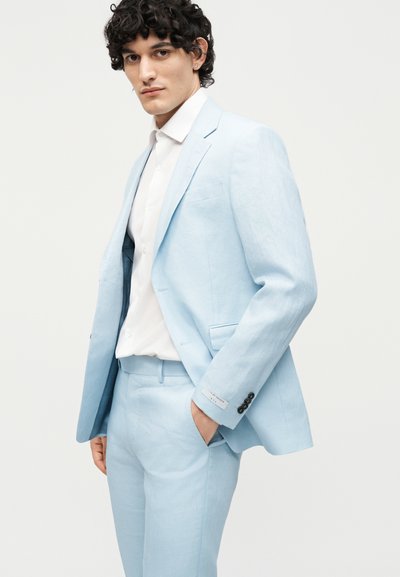 Νέος άνδρας με σγουρά μαλλιά, ντυμένος με ανοιχτό μπλε κοστούμι και λευκό πουκάμισο, στέκεται με το ένα χέρι στην τσέπη μπροστά σε απλό φόντο.