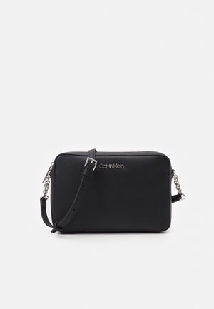 Zwarte rechthoekige Calvin Klein crossbodytas met verstelbare riem en zilveren kettingaccenten op een witte achtergrond.
