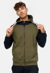 Sudadera con cremallera en color verde oliva con mangas en navy, capucha ajustable y bolsillos delanteros. Presenta un logo en el pecho y puños acanalados.