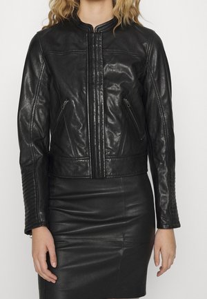 Mujer con chaqueta de cuero negra ajustada con acolchado vertical en las mangas y bolsillos laterales con cremallera, combinada con una falda de cuero negra.
