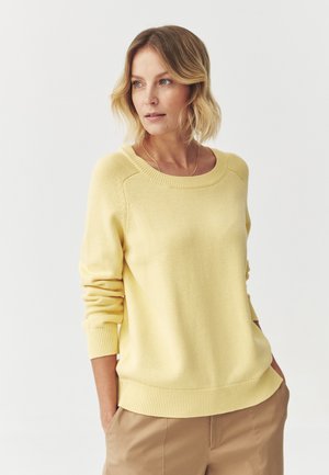 Pulover galben tricotat cu decolteu rotund, manșete și tiv cu nervuri, având o textură moale și o croială lejeră. Asortat cu pantaloni bej.
