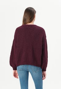 Oversized burgundský pletený svetr s texturovaným povrchem, spadlými rameny a žebrovanými manžetami. V kombinaci s úzkými modrými džíny.