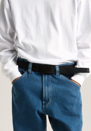 Schwarzer Stoffgürtel mit einer rechteckigen schwarzen Schnalle, getragen über blauen Jeans und einem weißen Langarmhemd.