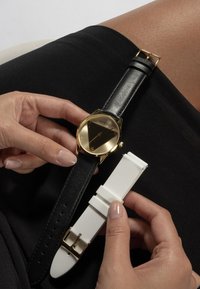 Montre dorée et noire avec un cadran triangulaire et un bracelet en cuir, près d'un bracelet de remplacement en silicone blanc. Des mains ajustent la montre.