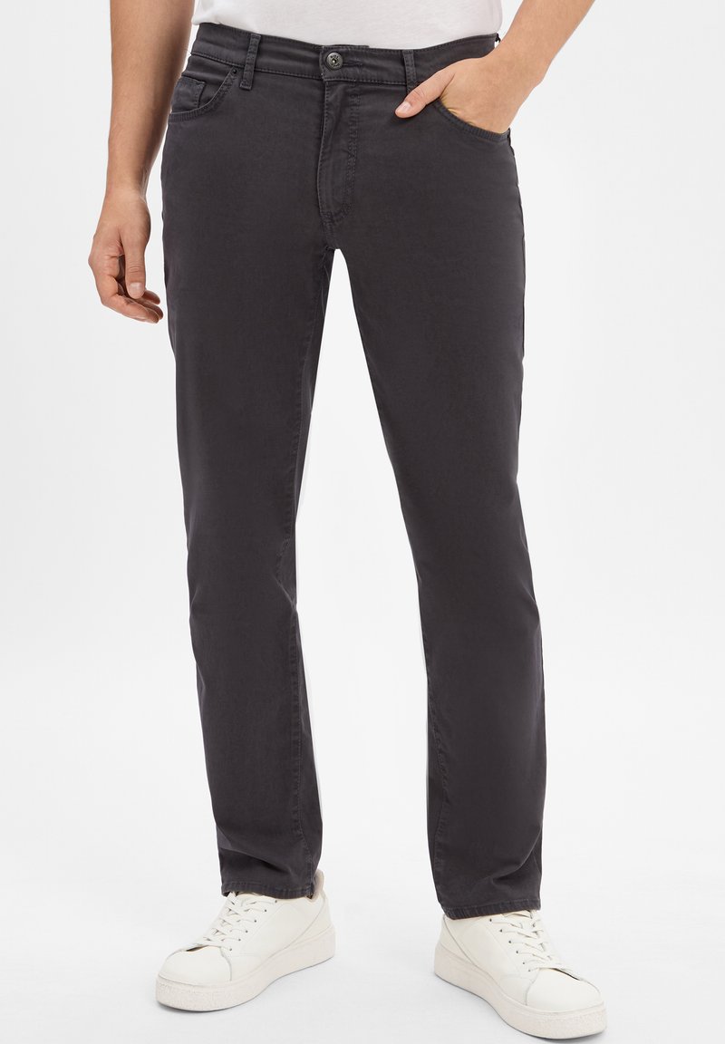 Uomo che indossa pantaloni dritti grigio scuro con sneakers bianche, una mano in tasca, in piedi contro uno sfondo bianco.