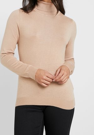 Femme portant un pull à col roulé beige et un pantalon noir, les mains jointes devant un fond uni.
