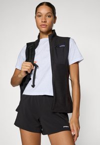 Zwarte, gestructureerde vest met een ritszak op de borst, gedragen over een wit t-shirt en zwarte shorts. Het vest heeft verstelbare banden op de schouders.