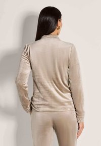 Vrouw met lang, donker haar die een beige fluwelen longsleeve shirt en bijpassende broek draagt, gezien van achteren met de rechterhand in de zak.