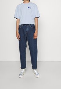 Jasnoniebieska bawełniana koszulka z graficznym wzorem fali na piersi, w zestawie z ciemnoniebieskimi dżinsami. Całość dopełniają białe sneakersy za kostkę.