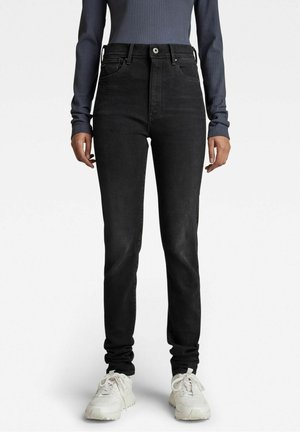 Femme portant un jean noir taille haute, un haut à manches longues gris côtelé et des baskets blanches, se tenant devant un fond uni.