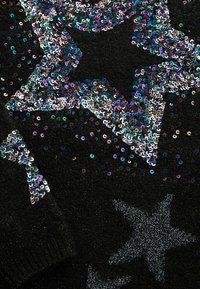 Pull noir orné de paillettes en forme d'étoiles aux teintes argentées et irisées, sur un tissu à texture apparente.