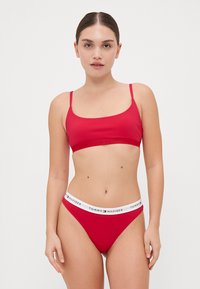 Rød bikini-sett med en tilpasset bralette-top med tynne stropper og matchende thong-bunn, begge med en logo-livlinje. Glatt tekstur.