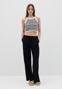 Top de tirantes recortado en color crema con rayas negras en forma de ola, combinado con pantalones holgados negros con cordón. La modelo está de pie con las manos en los bolsillos.