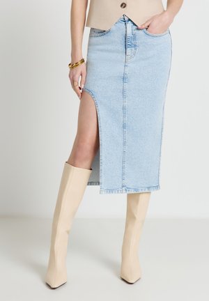 Gonna midi in denim azzurro chiaro con spacco alto laterale, indossata con stivali beige a punta al ginocchio e braccialetti dorati.