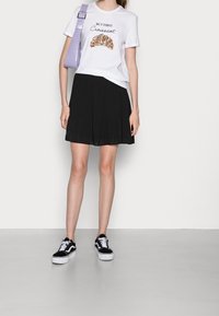 Vit t-shirt med grafisk text och en design av en croissant, kombinerad med en svart plisserad kjol. Svart och vita sneakers fulländar outfiten.