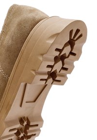 Gros plan sur la semelle d'une botte en daim beige montrant un motif de semelle profond et agressif, conçu pour l'adhérence et la durabilité.